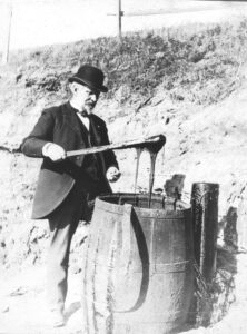 Black and white photo of a gentleman using a long stick to pull tar from a barrel.
