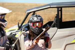 State Park Off Highway Vehicles (OHV) interpreter stands in front of a side-by-side vehicle wearing a black helmet, red mirrored googles, and holding an incident report on a clipboard.